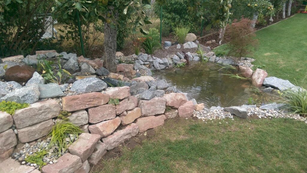 Gartengestaltung: Gartenanlage mit Teich, Naturstein-Mauer, Pflanzungen und Rasen